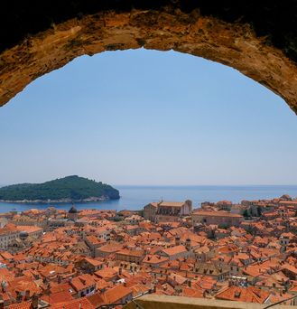 Blick durch einen Torbogen auf ein Häusermeer mit orangenen Dächern. Am Horizont das Meer und eine nahe gelegene Insel