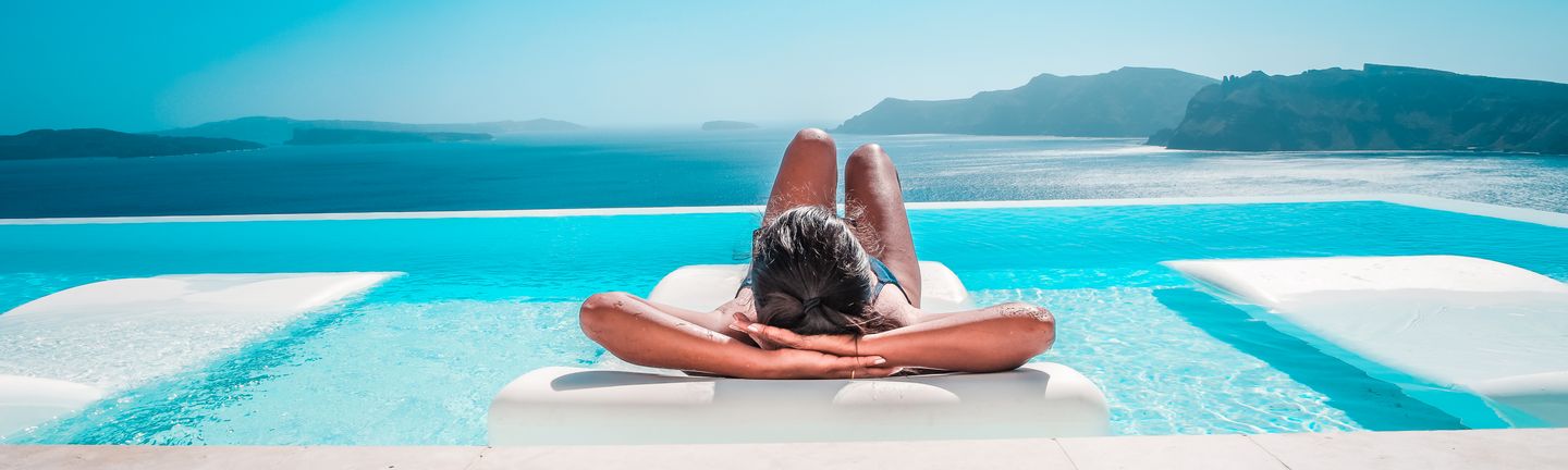 Eine braungebrannte junge Frau in Badekleidung, liegt entspannt auf einer weißen liege auf einem türkisen Infinitypool. Von diesem aus hat man einen unglaublichen Ausblick auf das Meer mit Bergen.