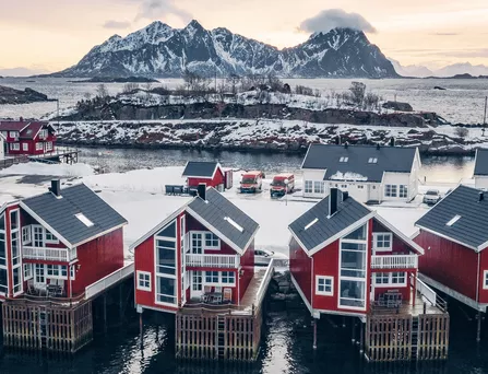 Rote Häuser stehen direkt am Ufer eines Gewässers, der Boden ist mit Schnee bedeckt im Hintergrund ein riesiger schneebedeckter Berg.