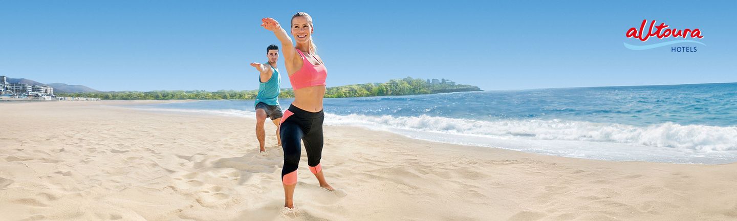 An einem hellen Sandstrand stehen eine Frau und ein Mann zwischen 25 und 35 in einer Yogapose und Sportklamotten und lächeln.