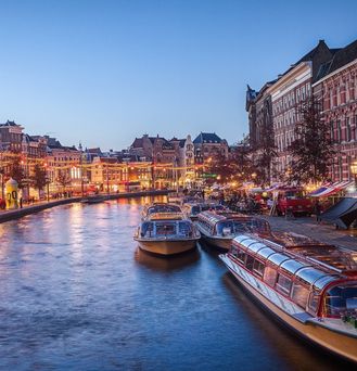 Eine Gracht in Amsterdam zeigt sich im Abendlicht. Das Ufer ist mit weihnachtlichen Lichtern beleuchtet.