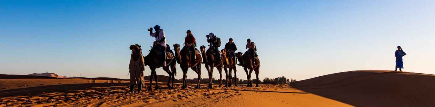 Eine Kamelkaravane aus 5 Kamelen und Touristen auf ihnen mit Ferngläsern lädt durch die Wüste.