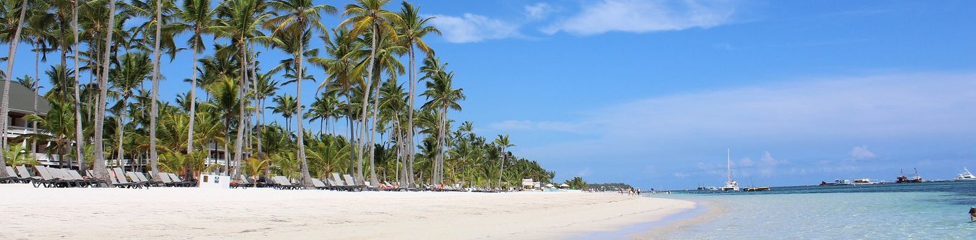 Urlaub in der Dom Rep: Sonnenliegen und Palmen am weißen Strand und seichtem Wasser, auf welchem einige Segelboote anliegen.