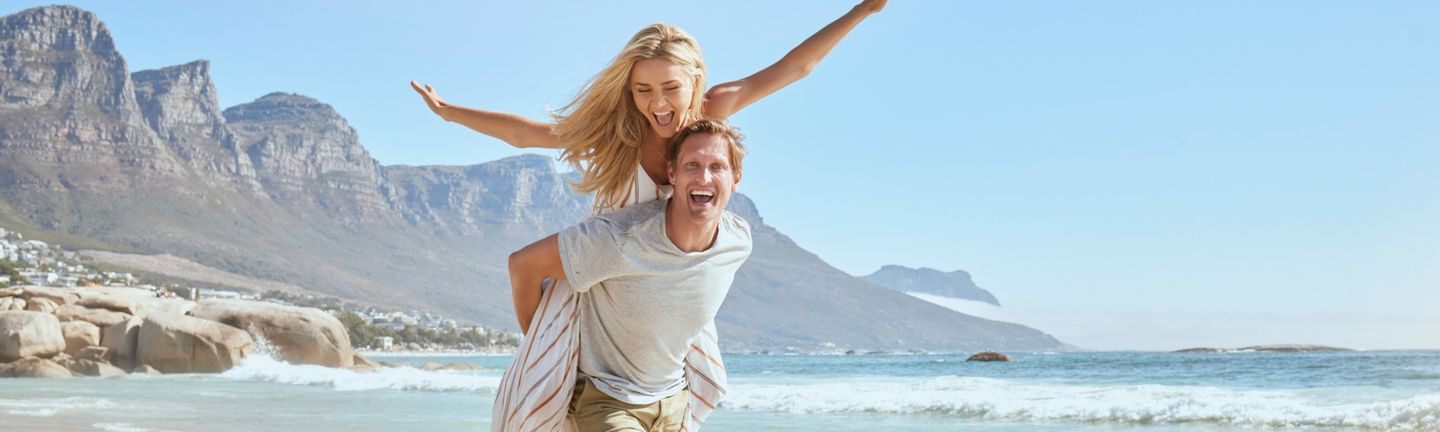 Reiseziele weltweit: Ein junges, lachendes Paar am Strand, im Hintergrund Berge