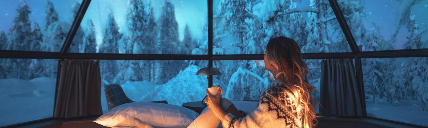 Eine Frau mit Norwegerpulli sitzt unter einer Glaskuppel in einer verschneiten Landschaft.