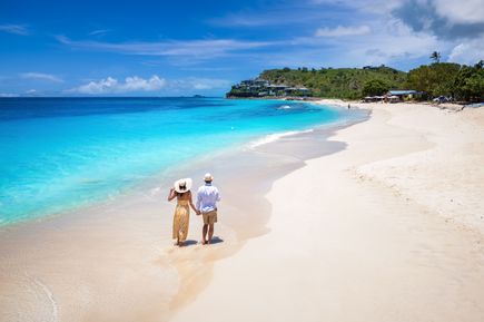 Ein Paar mit Sommerkleidung läuft Hand-in-Hand an einem weißen Sandstrand mit kristallklarem türkisen Wasser entlang.