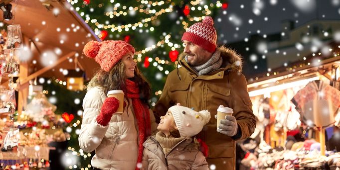 Junge Eltern stehen mit ihrer kleinen Tochter in Winterkleidung auf einem Weihnachtsmarkt. In der Luft glitzern die Schneeflocken und alle schauen sich mit einem To-go Becher in der Hand an.