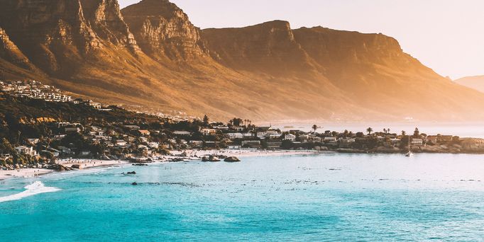 DAs blaue Meer grenzt an eine steile Bergkette, davor eine Stadt mit Palmen