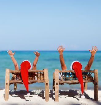 Zwei Urlauber mit Weihnachtsmannmützen liegen auf Holzliegestühlen am Strand und reißen die Arme in die Luft