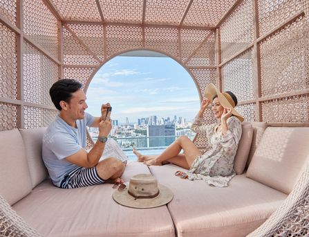 Ein asiatisches Pärchen sitzt in einer modernen Cabana und er schießt ein Foto von ihr. Durch ein rundes Loch ist die Skyline von Singapur zu sehen.