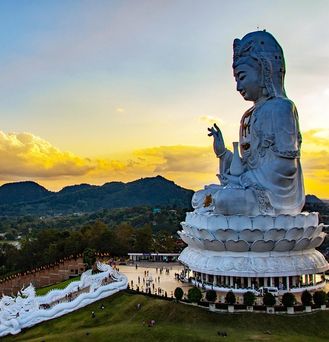 Eine riesige weiße Buddah Statue in einer grünen Landschaft unter orange-gelbem Himmel.