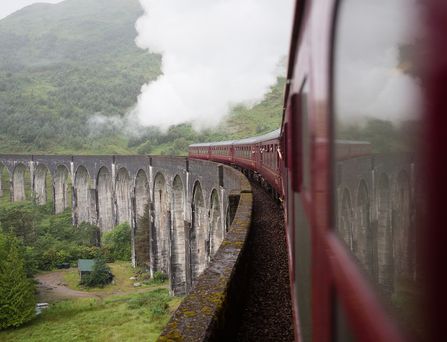 Ein roter Zug fährt über eine steinerne Brücke in einer grünen Landschaft.