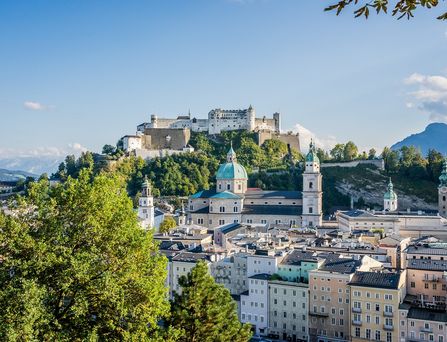 Auf einem Berg befindet sich eine historische Burg und im Tal erstreckt sich die Salzburger Skyline mit vielen hübschen, historischen Gebäuden. Im Hintergrund die Berglandschaft und die Stadt wird immer wieder von Bäumen durchzogen.