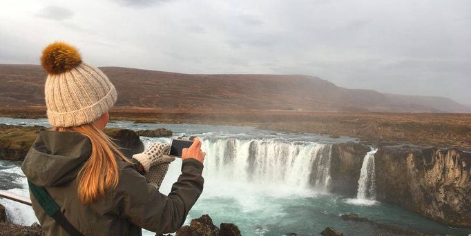 Eine blonde Frau mit Pudelmütze steht an einem Abhang mit dem Rücken zum Zuschauer. Sie fotografiert einen Wasserfall, mit einer Berglandschaft im Hintergrund