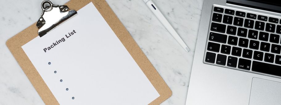 Ein Klemmbrett mit einer leeren Liste mit der Überschrift "packing list". Daneben liegt ein Stift und ein aufgeklappter Laptop.