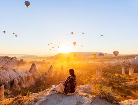 Eine Frau sitzt an einem Felsabsprung und blickt auf die Schlucht von Kappadokien. DIe Sonne geht gerade unter und taucht das gesamte Bild in ein orangenes Licht. Unzählige Heißluftballons schweben in der Luft.