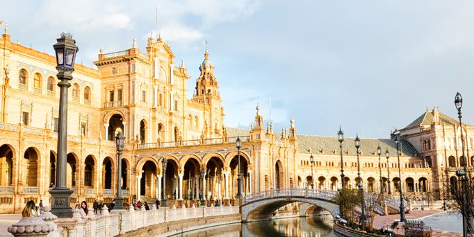 Die gold-gelbe Fassade eines prächtigen Bauwerks, vor einer Brücke, die über einen kleinen Fluss führt. Menschenleere Straßen.