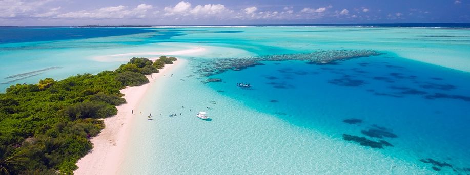 Ein schmaler weißer Sandstrand mit tropischr Vegetation, direkt am türkis, blauen Wasser mit zwei Booten am Ufer und einigen Menschen.