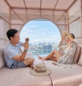 Ein asiatisches Pärchen sitzt in einer modernen Cabana und er schießt ein Foto von ihr. Durch ein rundes Loch ist die Skyline von Singapur zu sehen.