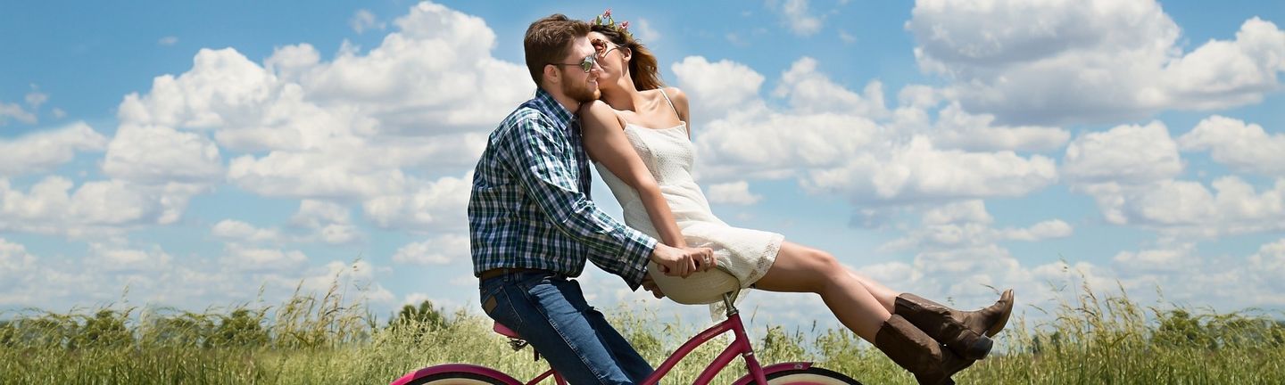 Kurzurlaub Valentinstag: Ein Mann fährt mit einem roten Fahrrad durch eine grüne Landschaft und eine Frau sitzt im weißen Sommerkleid auf dem Lenker