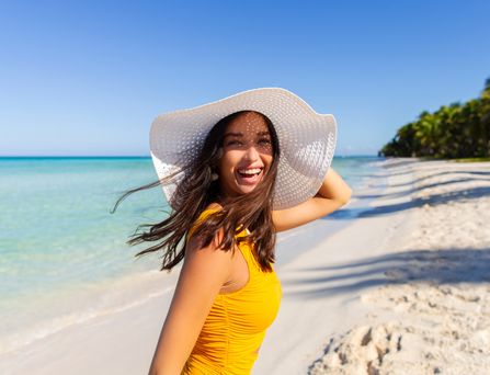 Eine Frau mit großem, weißen Sonnenhut läuft lachend an einem paradiesischen Strand entlang.