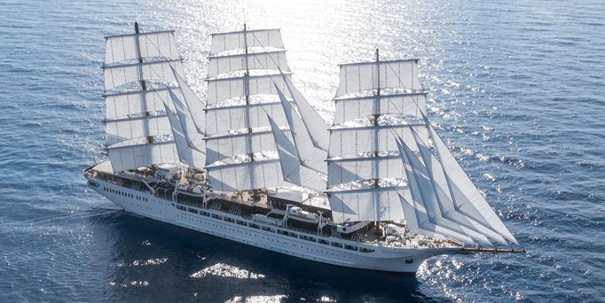 Ein großes 3-Masten Segelschiff gleitet über das tiefblaue, leicht wellige Wasser. Die Sonne glitzert auf der Wasseroberfläche.