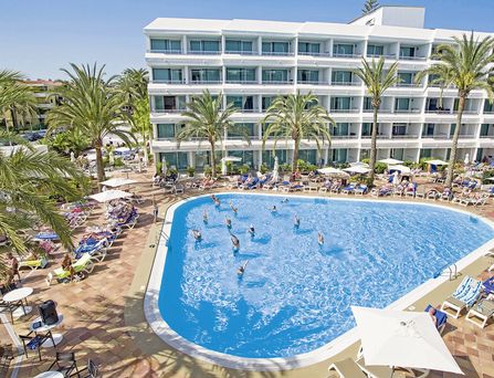 Ein ovalförmiger Pool befindet sich vor einem mehrstöckigen Gebäude umgeben von Palmen. Im Pool stehen Menschen und machen Gymnastikübungen.
