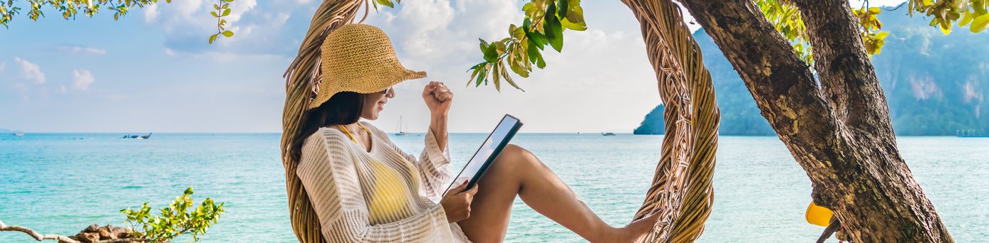 Frau sitzt entspannt im runden Bambushängesessel und liest auf einem Tablet. Im Hintergrund Meer.