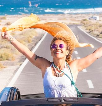 Eine sommerlich, hippimäßig gekleidete Frau sitzt in einem Cabrio mit offenem Dach. Sie steht im Auto und reist die Arme lachend nach oben und ein orangenes Tuch weht im Wind. Im Hintergrund das wilde Meer.