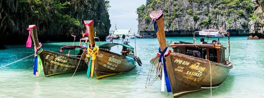 Drei typische thailändisch geschmückte Longtail Holzboote liegen in einer Bucht an. Im Hintergrund gigantische Kalksteinfelsen.