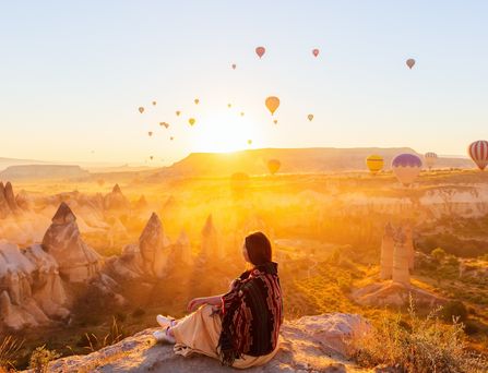 Eine junge Frau sitzt inmitten einer bizarren Berglandschaft und blickt in die Ferne. Die Sonne geht gleich hinter einem Berg unter und taucht die Szene in ein warmgelbes Licht. Der Himmel ist übersäht von unzähligen Heißluftballons.