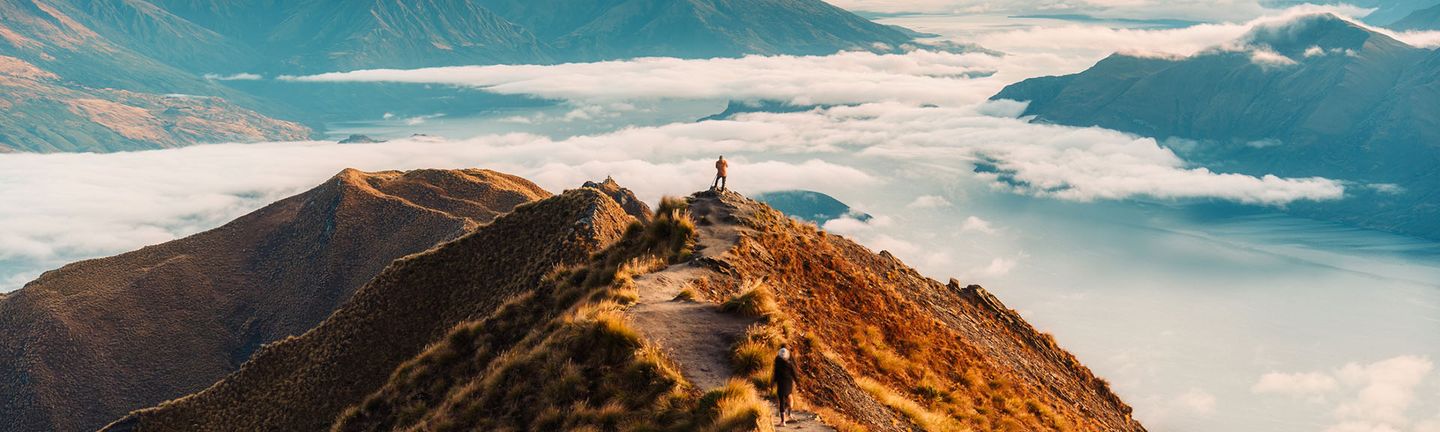 4 Personen laufen auf dem Gipfel eines braunen Berges der hoch über den Wolken tront