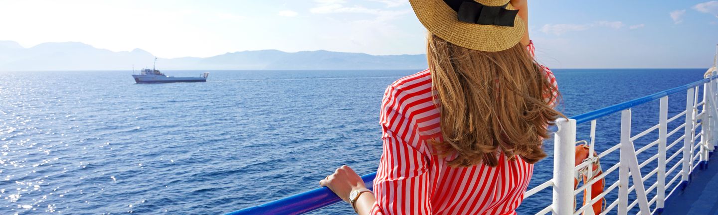 Eine Frau in sommerlicher Kleidung steht am Geländer eines Kreuzfahrtschiffes und blickt auf das offene, tiefblaue Meer. Darauf befindet sich ein kleineres Boot und im Hintergrund zeichnen sich Berge am Horizont ab.
