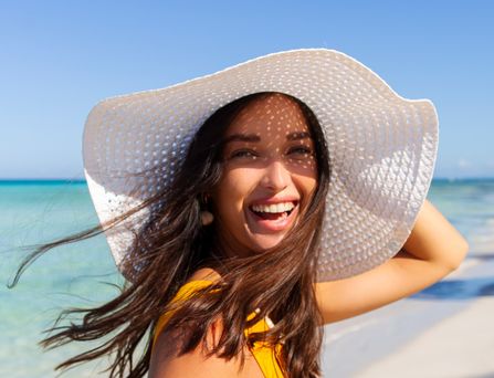 Eine Frau mit großem, weißen Sonnenhut läuft lachend an einem paradiesischen Strand entlang.