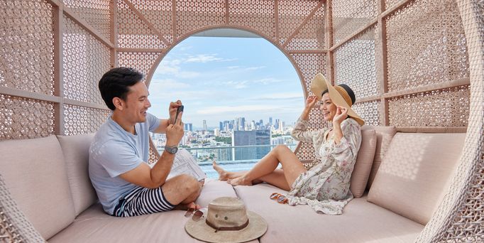 Ein asiatisches Pärchen sitzt in einer modernen Cabana und er schießt ein Foto von ihr. Durch ein rundes Loch ist die Skyline von Singapur zu sehen.