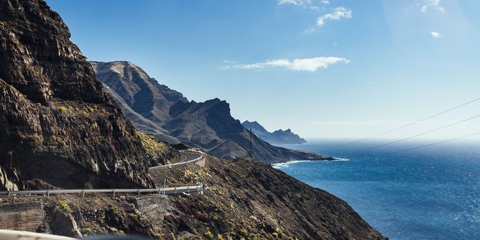 Am Rand einer steilen Schlucht führt eine kurvige Straße direkt am Meer entlang. Unten links befindet sich der rechte Außenspiegel eines Autos.