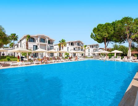 Um einen großen türkisen Pool stehen weiße mehrstöckige Häuser mit Palmen und Sonnenliegen davor.