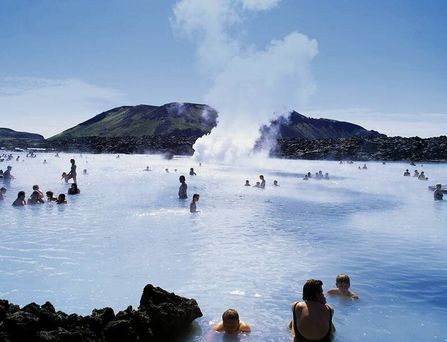 Viele Menschen baden in heißen Quellen aus denen Wasserdampf hochsteigt. Umgeben von Bergen und Felsen.
