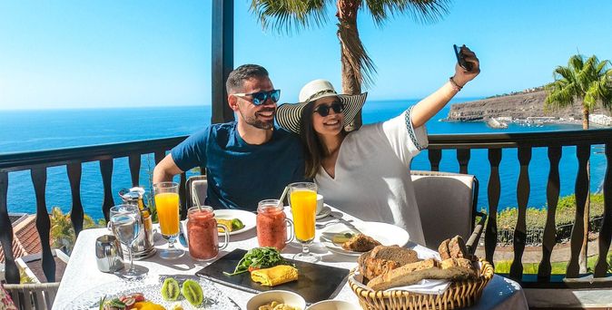 Ein junges Paar mit Sonnenbrillen und Sommerkleidung sitzt an einem reich gedeckten Frühstückstisch. Die Frau hält ihr Handy in die Luft und sie macht ein Selfie. Im Hintergrund das tiefblaue Meer und eine Palme.