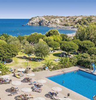 Direkt an einer felsigen Bucht am Meer liegt eine Wiese mit grünen Bäumen und ein Pool mit vielen Sonnenliegen mit Schirmen.