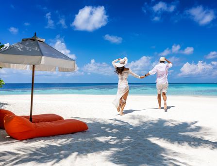 Hochzeitsreisen auf den Malediven: Ein Paar in weißer Kleidung läuft Händchen haltend einen weißen Strand entlang