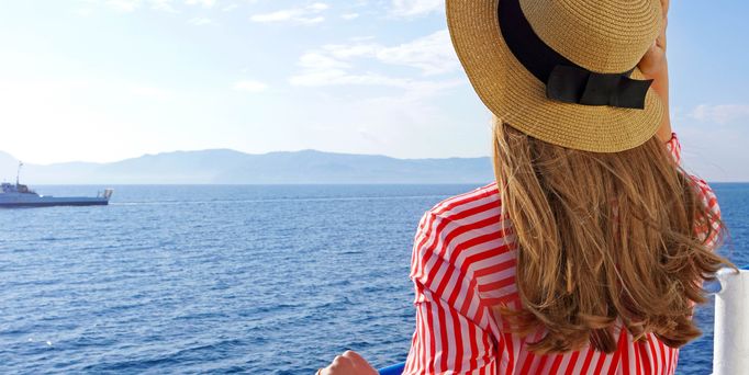 Reiseziele weltweit: Eine Frau mit Sonnenhut steht an der Rehling eines Kreuzfahrtschiffs und blickt aufs Meer