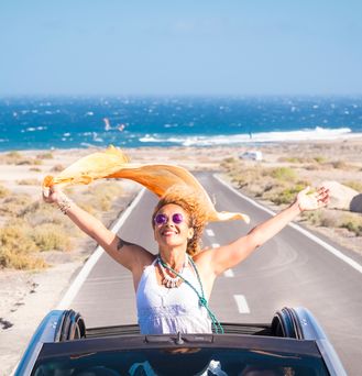 Eine sommerlich, hippimäßig gekleidete Frau sitzt in einem Cabrio mit offenem Dach. Sie steht im Auto und reist die Arme lachend nach oben und ein orangenes Tuch weht im Wind. Im Hintergrund das wilde Meer.