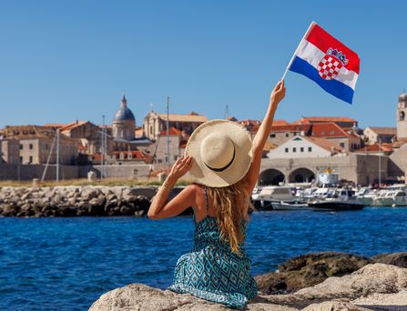 Eine junge Frau mit Sonnenhut und Kleid sitzt auf einem Felsen am Hafen und blickt auf das tiefblaue Wasser und die gegenüberliegende Stadt. Sie hält eine kroatische Flagge in der rechten Hand und hält sie hoch.