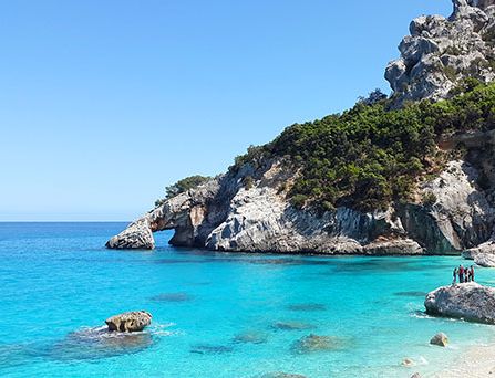 Kristallklares, türkises Wasser an einer steinigen, steilen Bucht. Ein paar Menschen stehen auf einem Felsen.