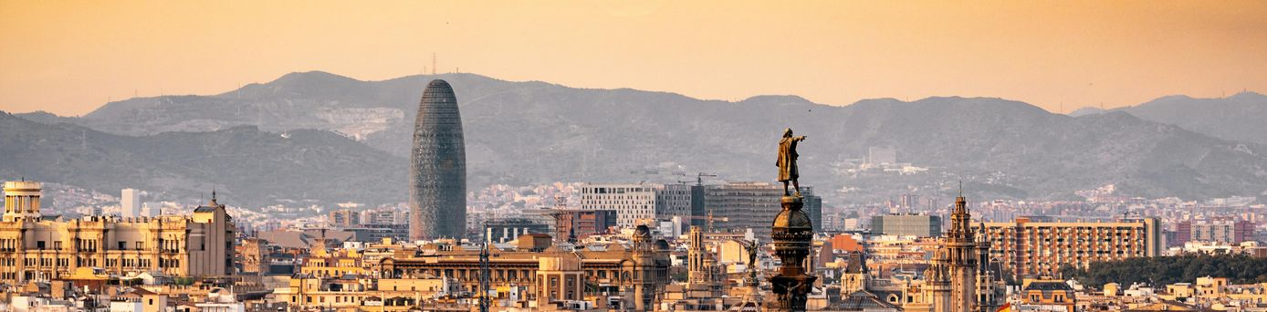 Die Skyline einer spanischen Stadt erstreckt sich in gold, gelbem Licht. Im Hintergrund sind dunkle, große Berge und darüber die runde, untergehende Sonne.