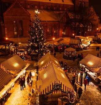 Auf einem dunklen Marktplatz mit bleuchtetem Weihnachtsbaum stehen Weihnachtsmarkthütten. Im Hintergrund befindet sich eine Kirche