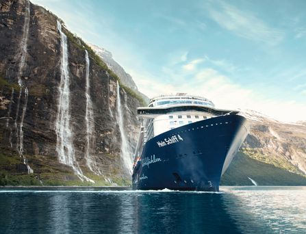 Das blau-weiße Kreuzfahrtschiff "Mein Schiff 4" gleitet durch das dunkle Meer entlang eines steilen Felsens mit Wasserfall.