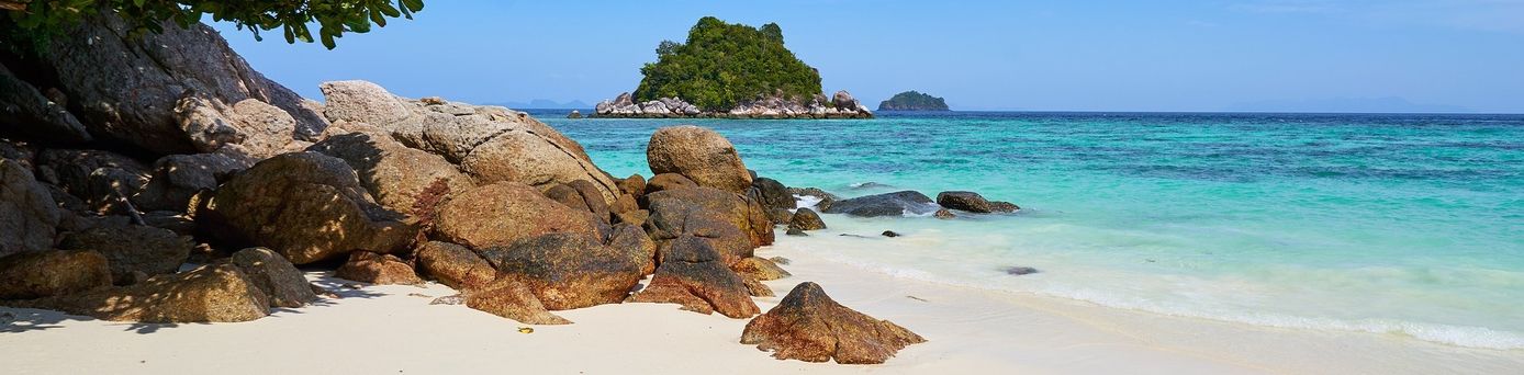 Ein feinsandiger, weißer Strand mit großen Steinen, welche ins türkisblaue, klare Meer ragen. Im Hintergrund eine kleine Insel.