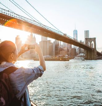 Eine Frau mit Rucksack und Smartphone in der Hand fotografiert die Brooklynbridge. Sie steht unten direkt am Flussufer. 